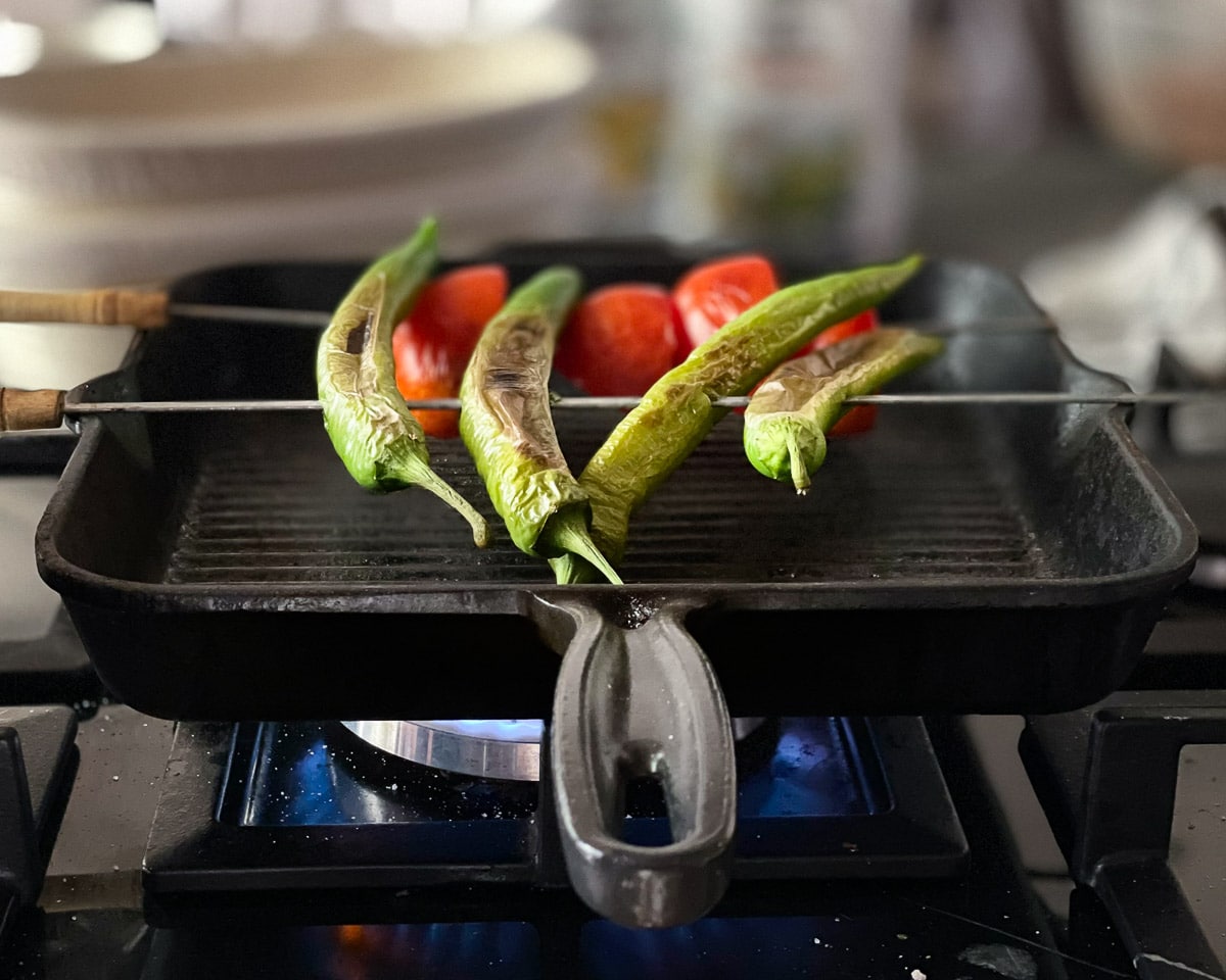 Paprika og tomat grilles på grillspyd liggende over en het grillpanne, med stor gassflamme synlige under. Det er mellomrom mellom bunnen av grillpana og grønnsakene, som  er tredd på grillspyd som hviler på kantene av grillpanna
