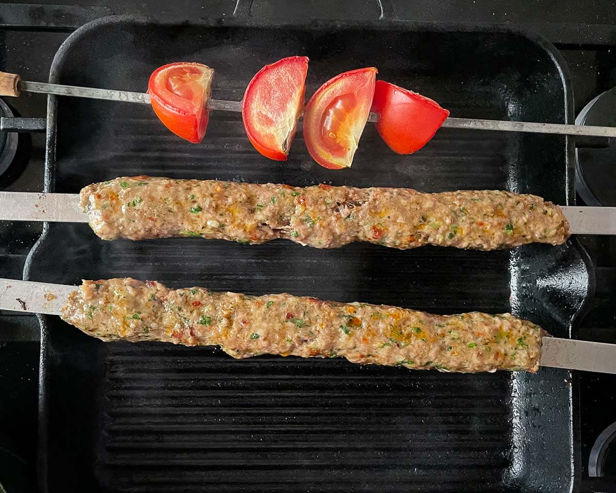 Adana-kebab grilles på flate grillspyd liggende over en het grillpanne, med stor gassflamme synlige under. Det er mellomrom mellom bunnen av grillpanna og kebaben, som er tredd på grillspyd som hviler på kantene av grillpanna